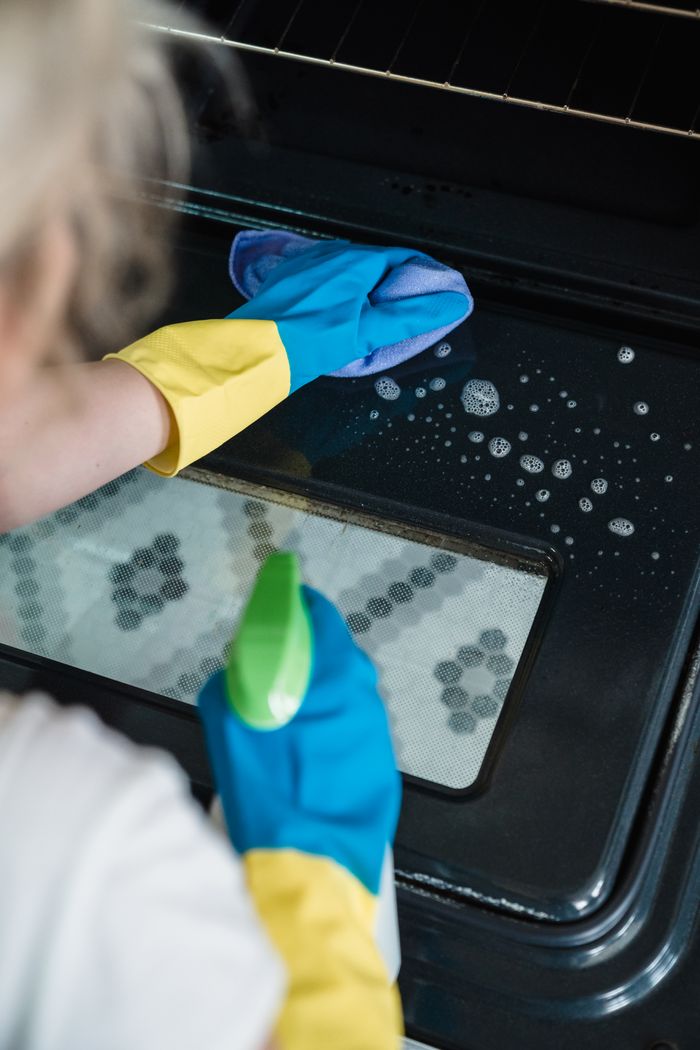 A person wearing gloves cleans an oven door using a spray and cloth.